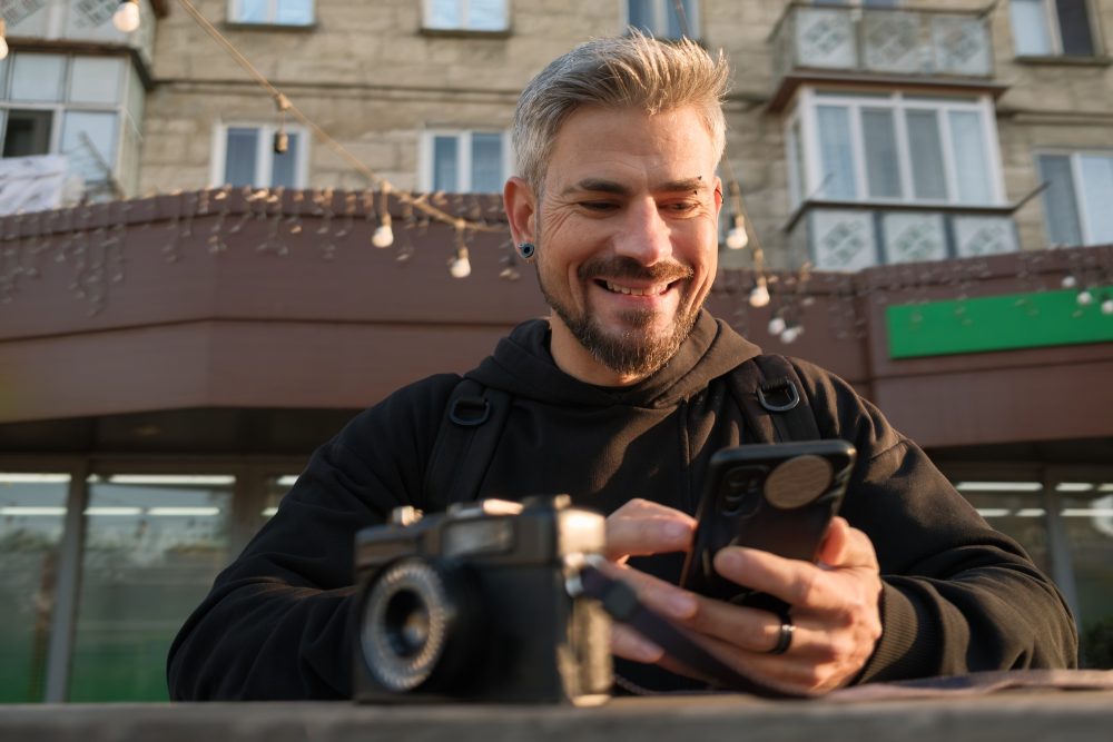 Man smiling while using phone with camera in sunny outdoor setting
